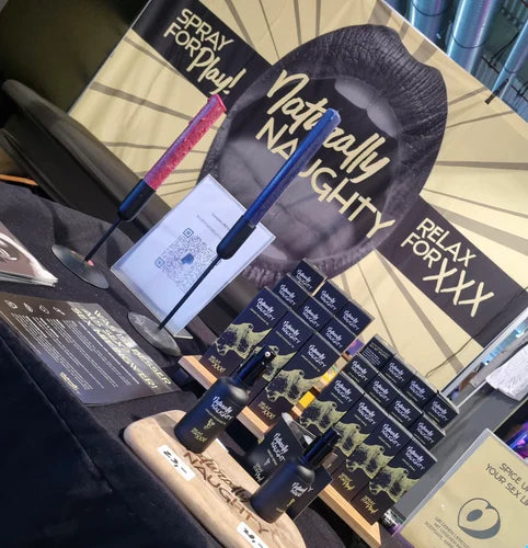 Messestand von Naturally Naughty mit Ingwerspray und -öl-Produkten, dekoriert mit Branding und auffälligem Banner mit dem Slogan 'Spray for Play' und 'Relax for XXX'.