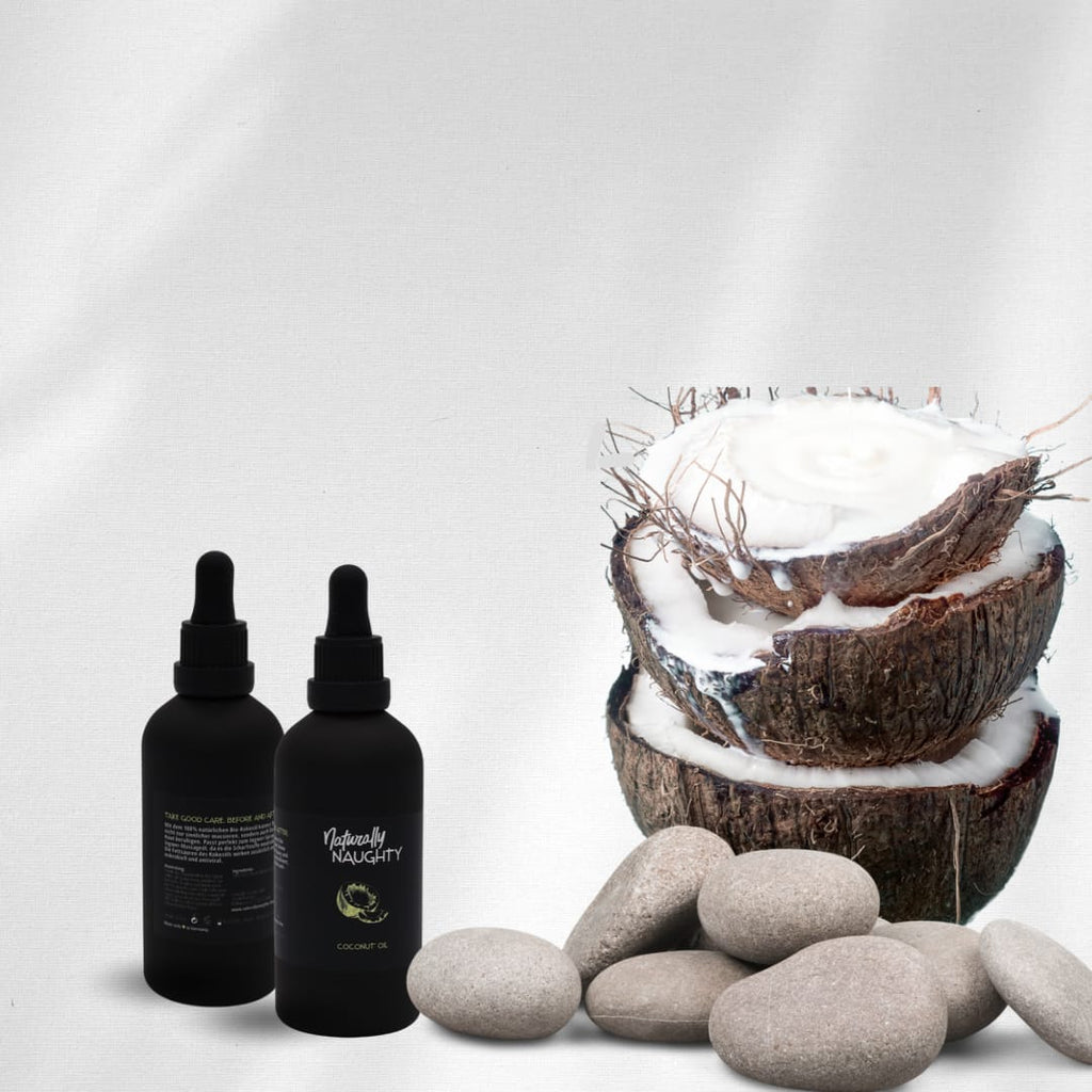 Natürliches Kokosnussöl von Naturally Naughty in zwei schwarzen Flaschen neben frischen, geöffneten Kokosnusshälften und glatten Steinen. Ideal für Massagen, Hautpflege und Wellness. Hochwertiges Bio-Kokosöl in edlem Design